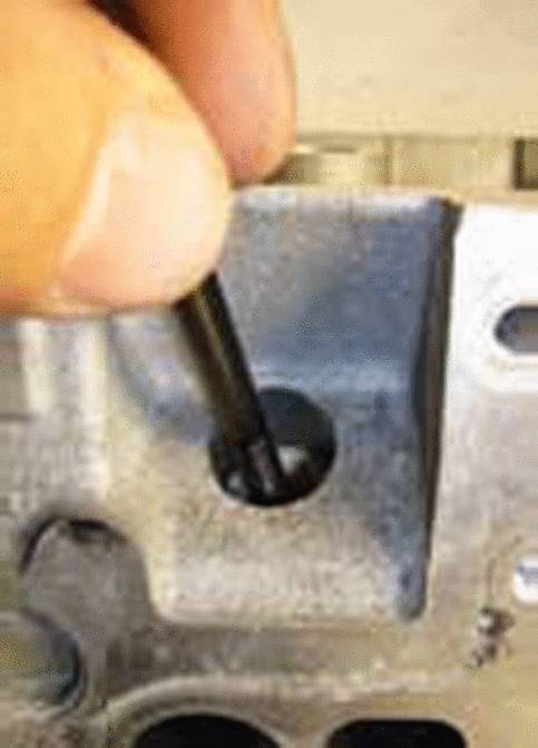 Eine Hand hält ein schwarzes Werkzeug und steckt es in ein Loch eines metallischen Bauteils.