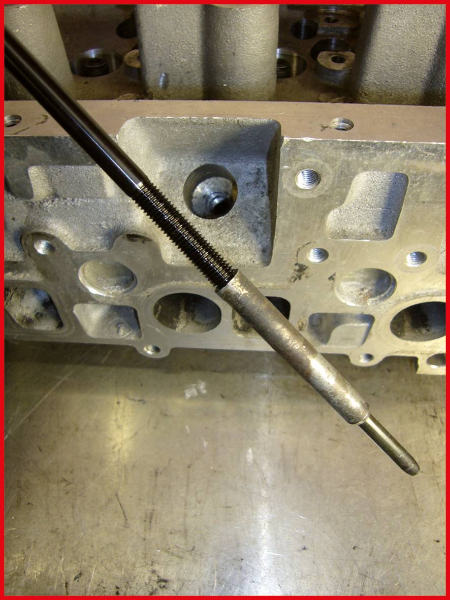 Ein beschädigter Zylinderkopf aus Aluminium mit einem abgebrochenen Bolzen, der herausragt. Hintergrund ist eine Werkstattfläche.