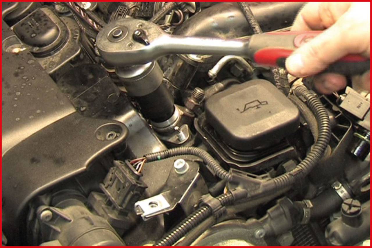 Eine Hand zieht mit einem Schraubenschlüssel eine Schraube an einem Autoteilmotor fest. Mehrere Kabel und Schläuche sind sichtbar.