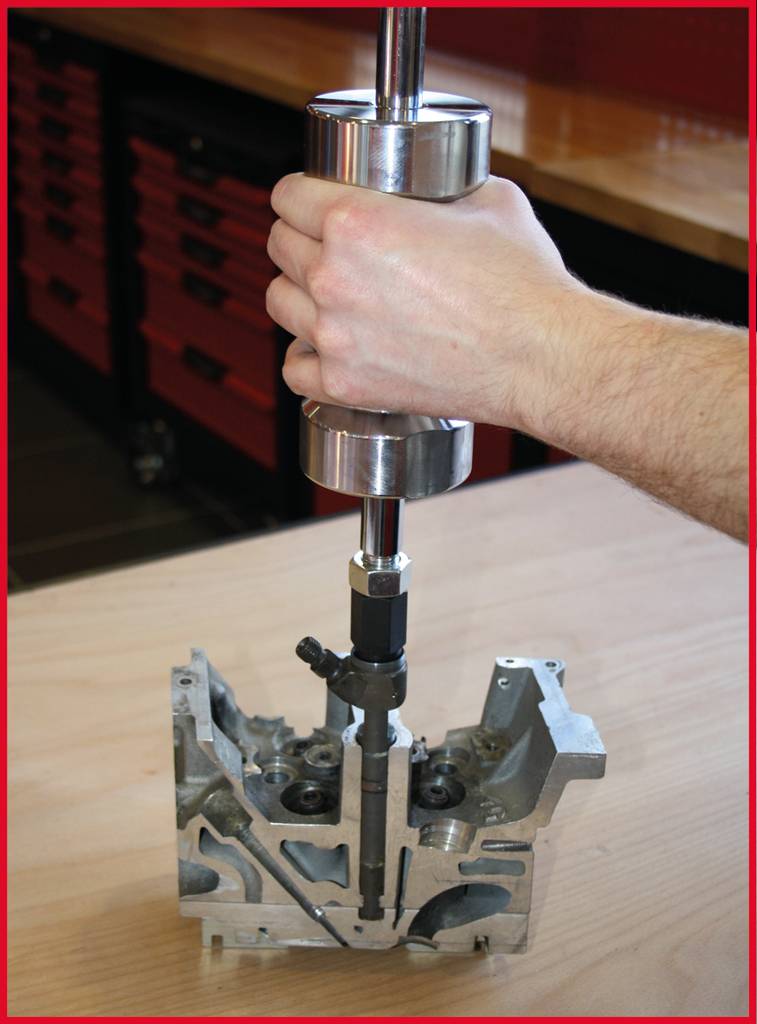 Ein Metallwerkzeug wird von einer Hand gehalten und in einen silbernen Motorteil eingeführt. Der Hintergrund zeigt eine Werkstatt.
