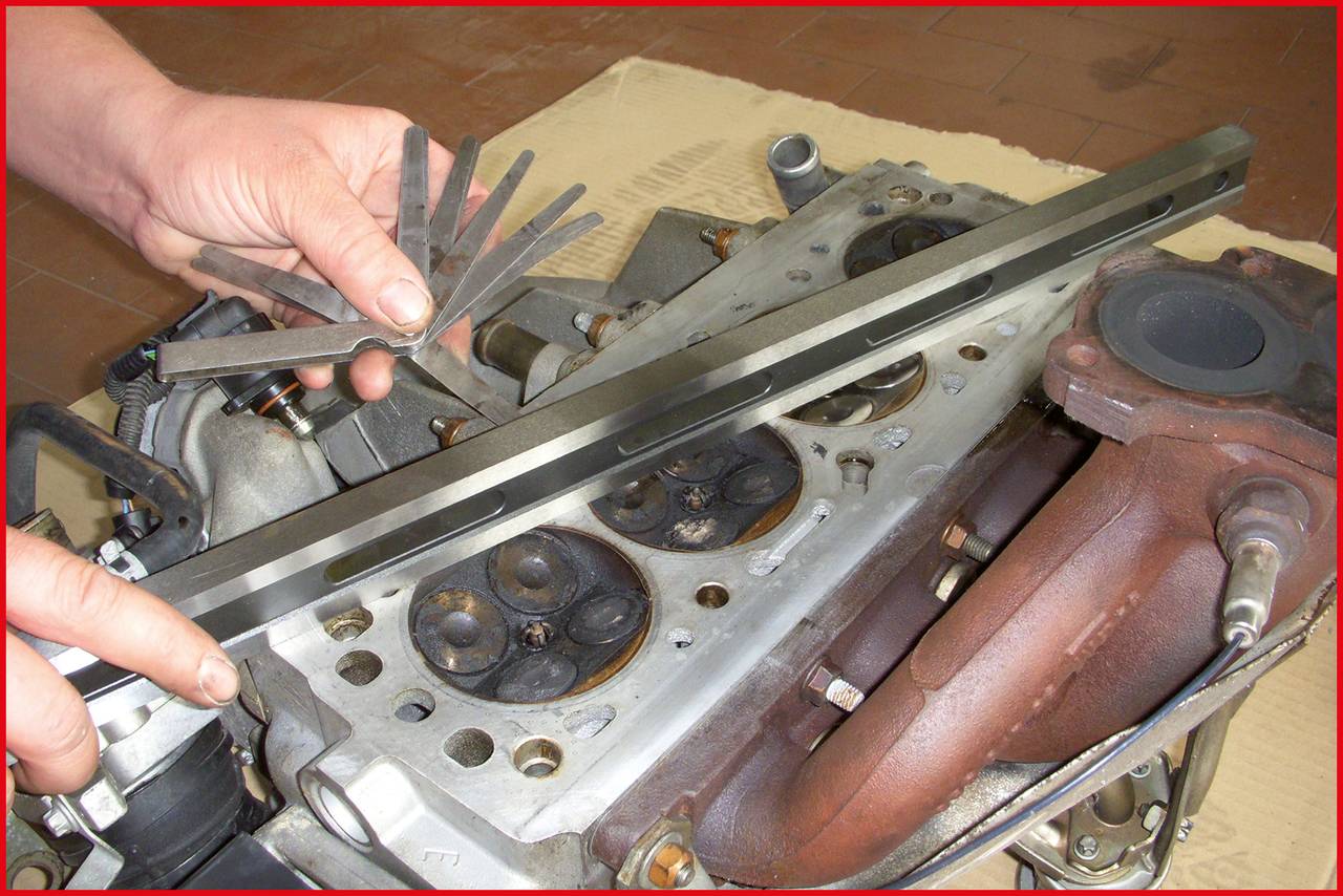 Eine Person misst den Ventilabstand eines Zylinderkopfes mit einer Fühlerlehre und einer geraden Kante bei der Motorreparatur.