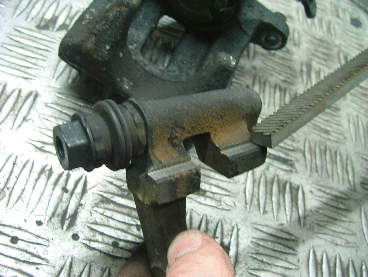 Nahaufnahme eines Bremssattels mit einer Feile auf einer Metalloberfläche. Eine Hand hält den Bremssattel in Position.