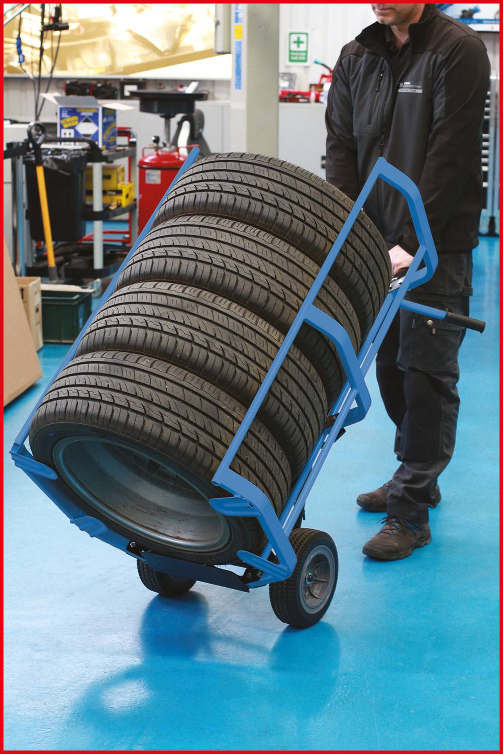 Mitarbeiter transportiert acht übereinandergestapelte Autoreifen mit einer blauen Sackkarre in einer Werkstatt.