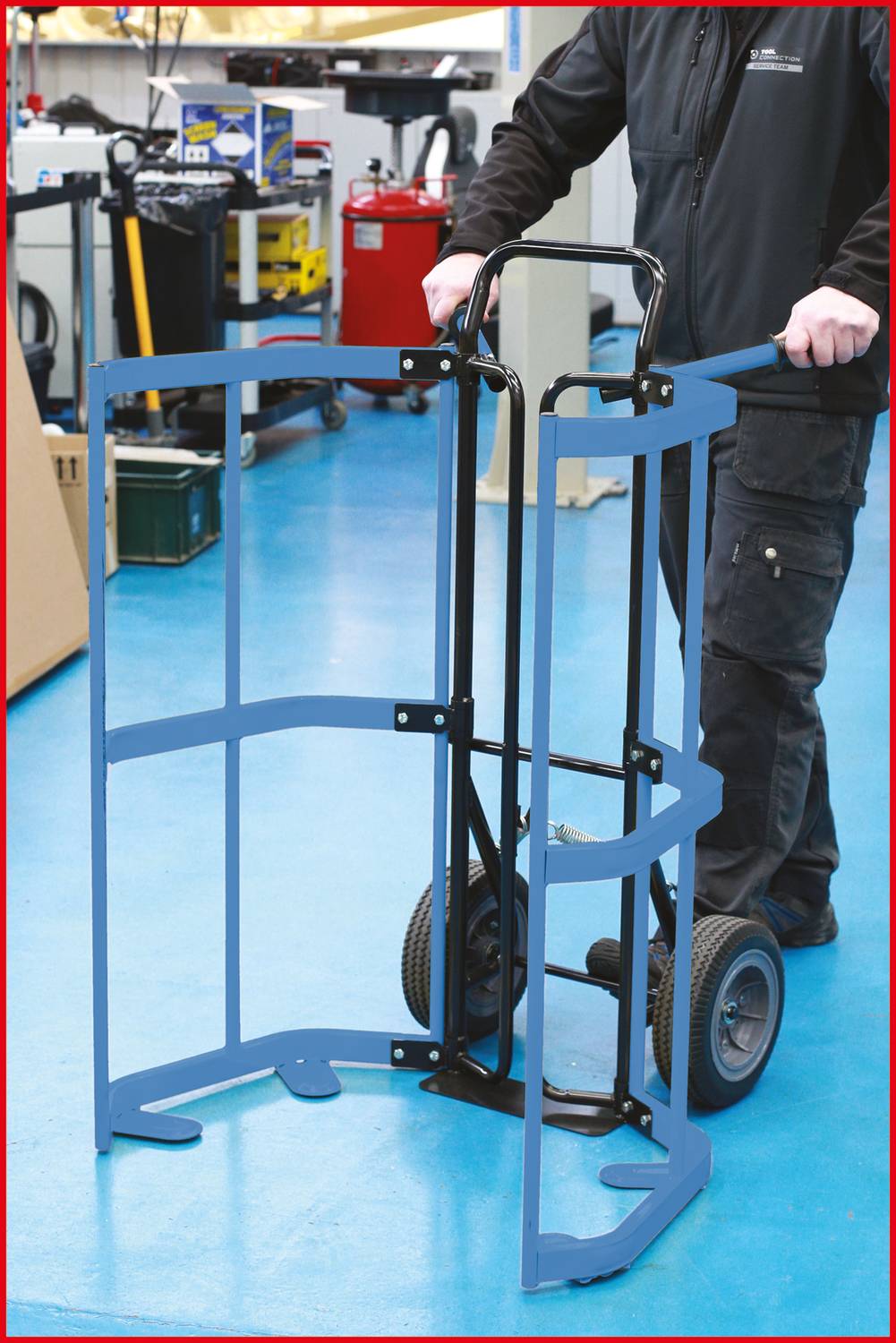 Ein Mann schiebt eine Sackkarre mit Rädern und einem blauen Schutzgestell in einer Werkstattumgebung. Werkzeug und Geräte im Hintergrund.