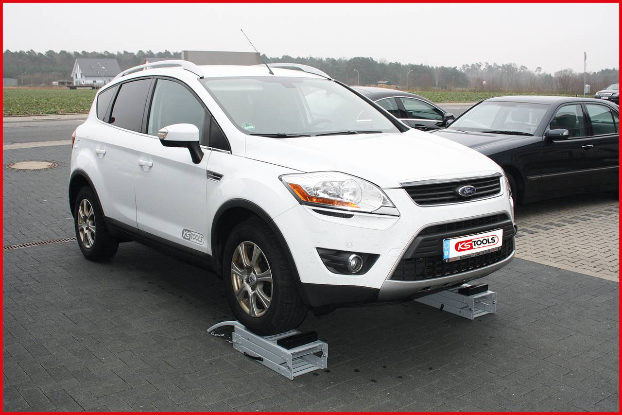 Ein weißes Auto steht auf einem mobilen Reifen-Wechsel-Helfer, umgeben von einem anderen Fahrzeug und einer ländlichen Straße im Hintergrund.