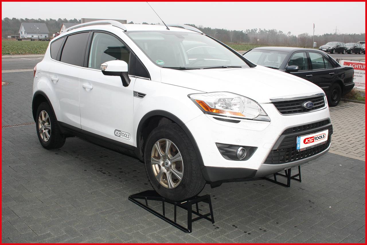 Ein weißes Auto steht erhoben auf Metallständern in einem freien Parkplatzbereich. Ein weiteres Auto ist im Hintergrund sichtbar.