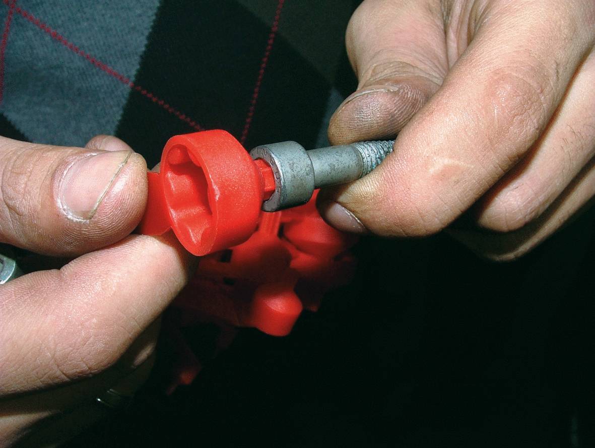 Nahaufnahme von Händen, die ein rotes Kunststoffteil und ein silbernes Metallwerkzeug zusammenfügen. Sie befinden sich in einem Reparatur- oder Montageszenario.