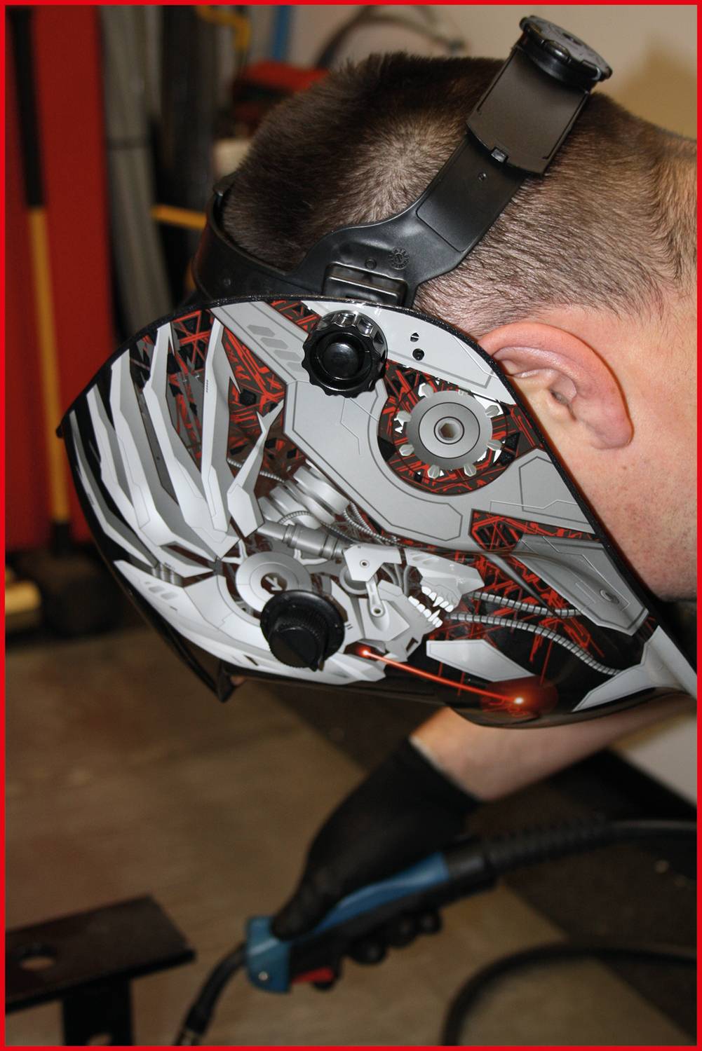 Eine Person trägt einen Schweißhelm mit modernem Design und hält ein Schweißgerät. Die Umgebung deutet auf eine Werkstatt hin.