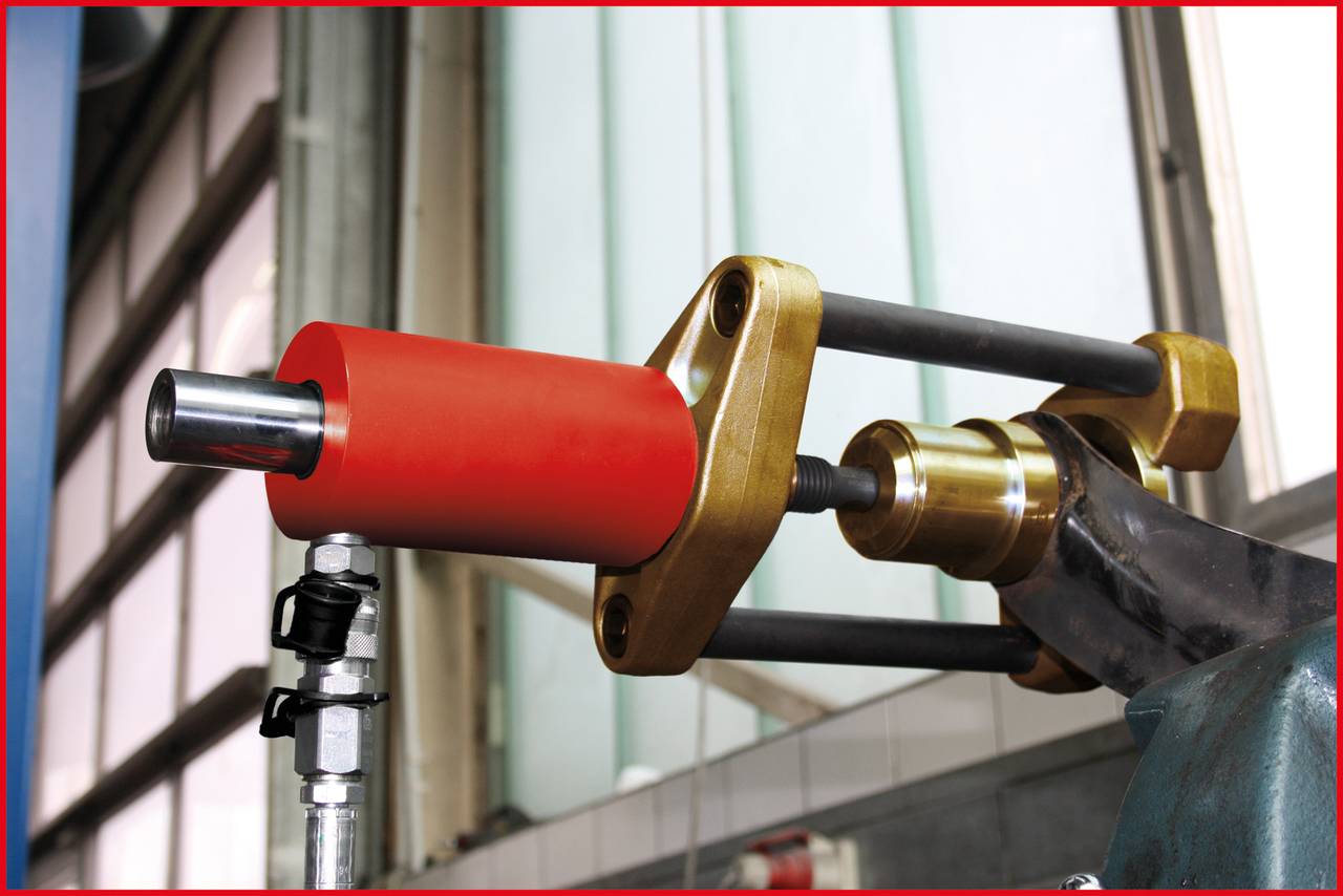 Eine Maschine mit einem roten zylinderförmigen Teil und goldenen Halterungen in einer Industrieumgebung. Fenster und Rohre im Hintergrund.