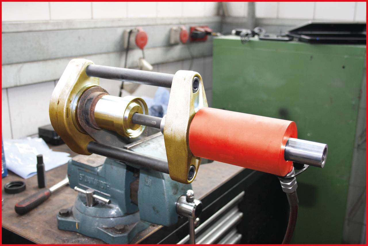Maschine in einer Werkstatt spannt ein Metallstück ein. Im Hintergrund sind Arbeitsmaterialien und ein grüner Schrank erkennbar.