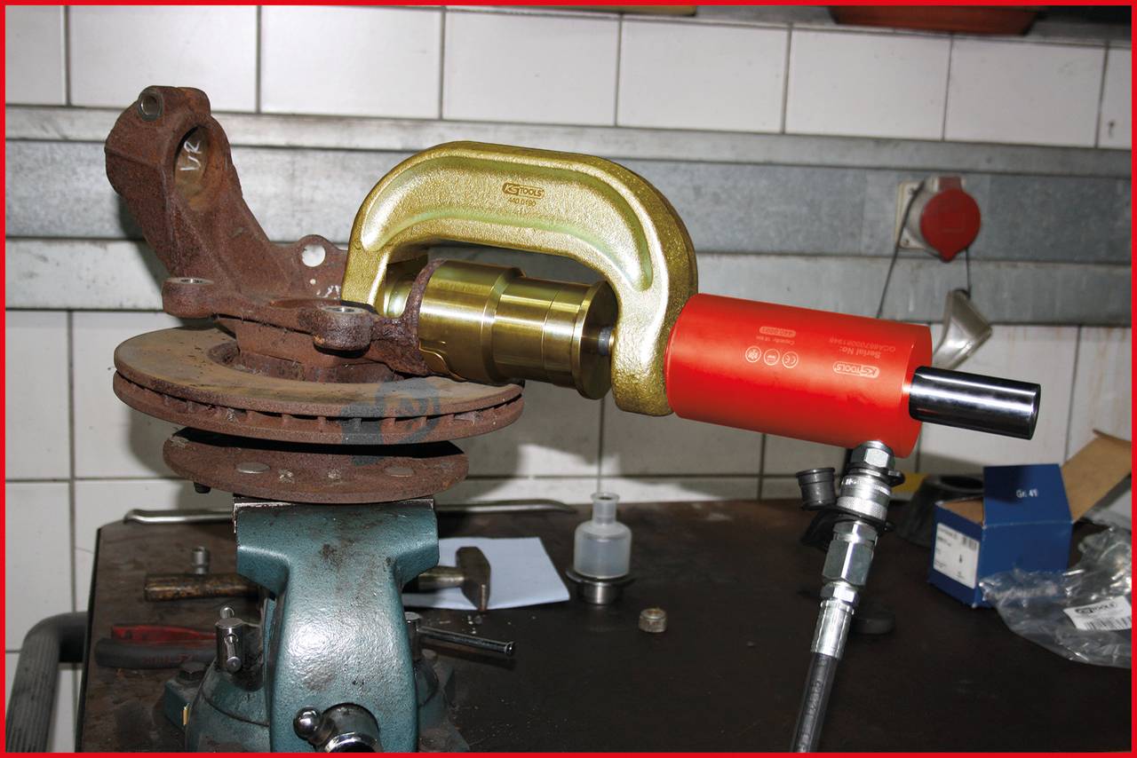 Bild eines rostigen Bremssattels in einer Werkstattklammer. Ein hydraulisches Werkzeug klemmt den Bremssattel, gezeigt auf einer Werkbank.