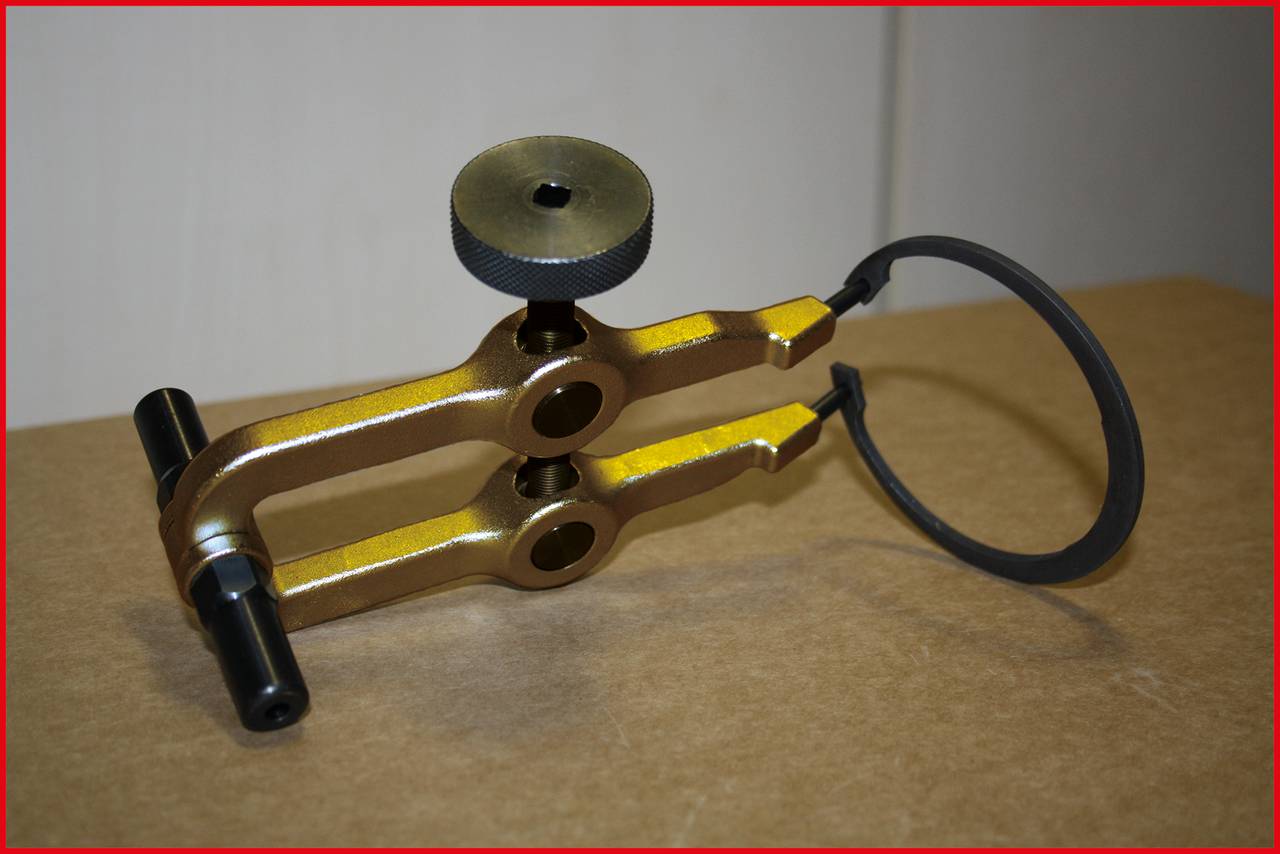 Metallisches Werkzeug auf Tisch platziert, mit Hebelmechanismus und schwarzem Rad. Es scheint für präzise mechanische Arbeiten gedacht zu sein.