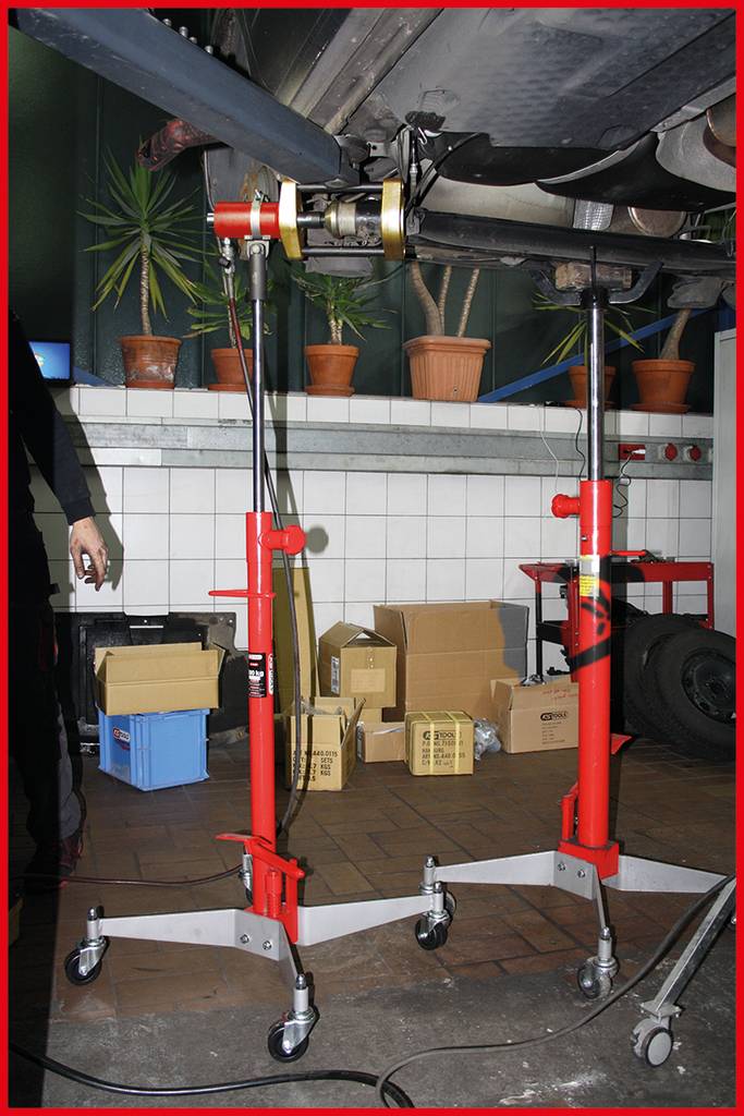 Ein rotes Fahrzeughebesystem hebt ein Autoteil in einer Werkstatt an. Im Hintergrund sind Pflanzentöpfe und Kartons sichtbar.