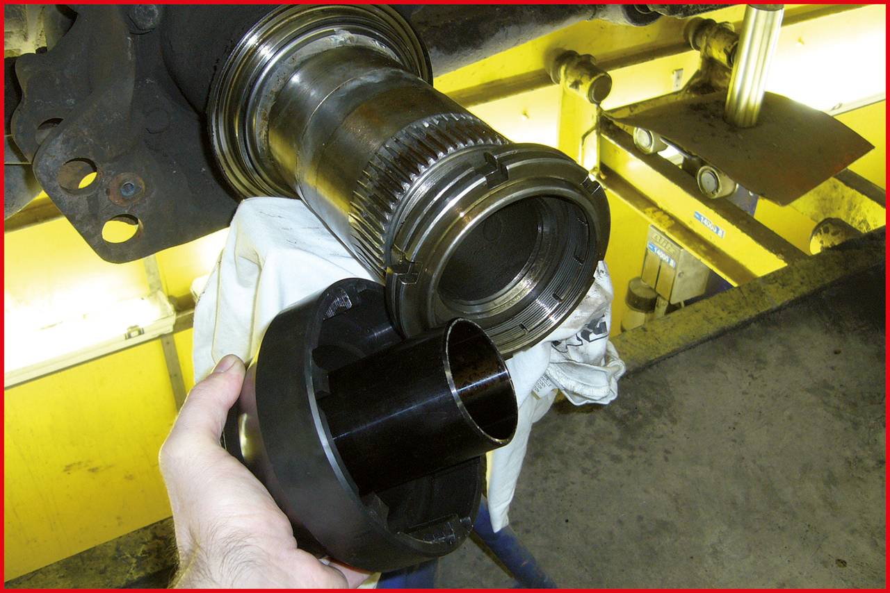 Eine Person hält ein schwarzes Metallteil vor eine Maschinenachse in einer Werkstatt. Hintergrund zeigt Werkzeuge und gelbe Maschinenoberfläche.