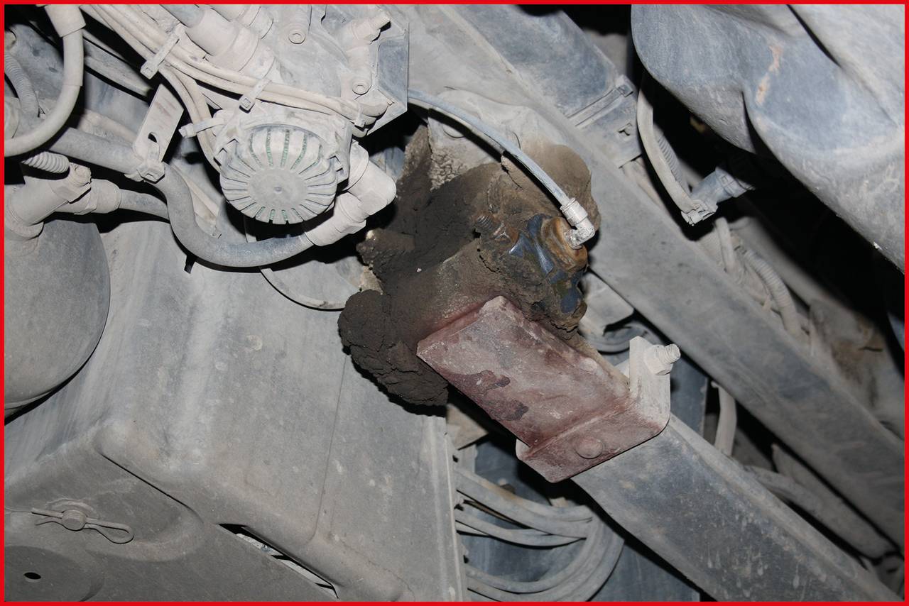 Eine Roststelle auf einem Fahrzeugunterboden, nahe einem mechanischen Bauteil, umgeben von Metallteilen und Kabeln.