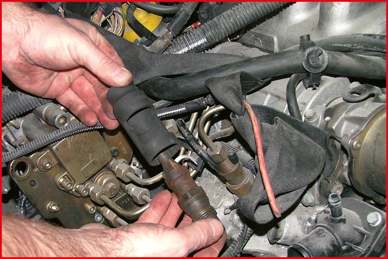 Eine Person hält eine Zündkerze über einem komplexen Motor mit sichtbaren Kabeln und Rohren. Die Szene zeigt eine Autoreparatur.