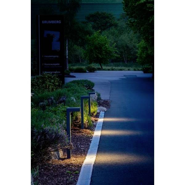 Straße in der Dämmerung mit beleuchteten Wegleuchten entlang eines Gehwegs. Wälder und Gräser im Hintergrund.