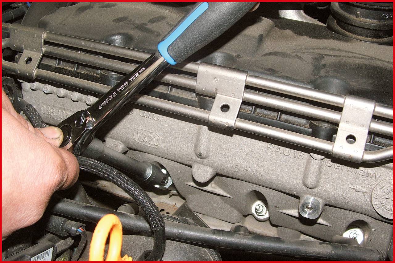 Eine Hand hält einen Schraubenschlüssel, der an einem Ventildeckel eines Automotors arbeitet. Umgebung zeigt verschiedene Motorenteile.