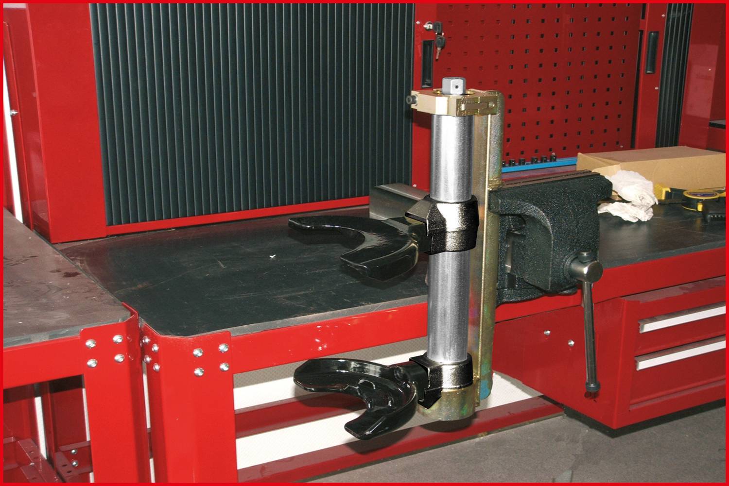 Eine Metallvorrichtung mit zwei Greifarmen ist auf einer Werkbank in einer Werkstatt montiert, umgeben von roten Schränken und Werkzeugen.