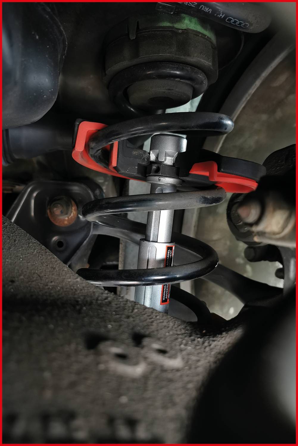 Nahaufnahme eines Auto-Stoßdämpfers mit Metallfeder und rotem Spannwerkzeug. Einbau im Radkasten zu sehen.