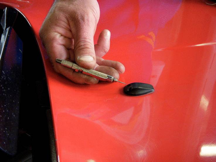 Eine Hand hält ein kleines Metallwerkzeug neben einem schwarzen Seitenspiegel auf einer glänzenden roten Autotür.
