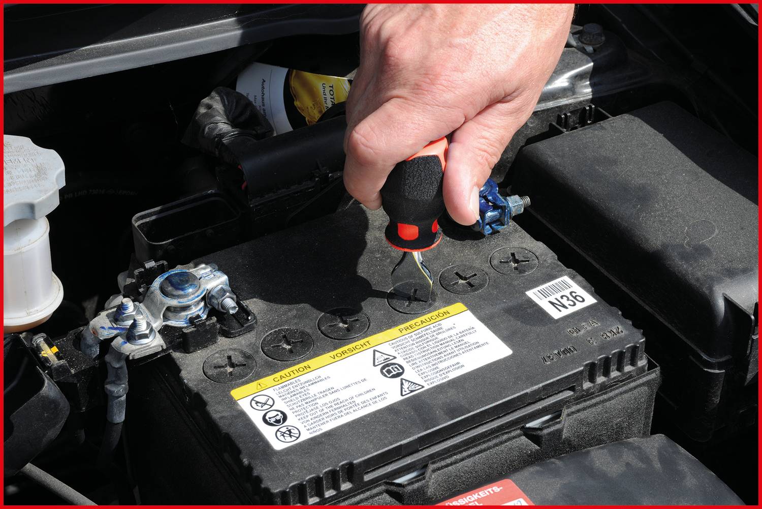 Hand prüft den Flüssigkeitsstand einer Autobatterie mit einem Schraubenzieher unter der Motorhaube. Batteriewarnhinweise sichtbar.