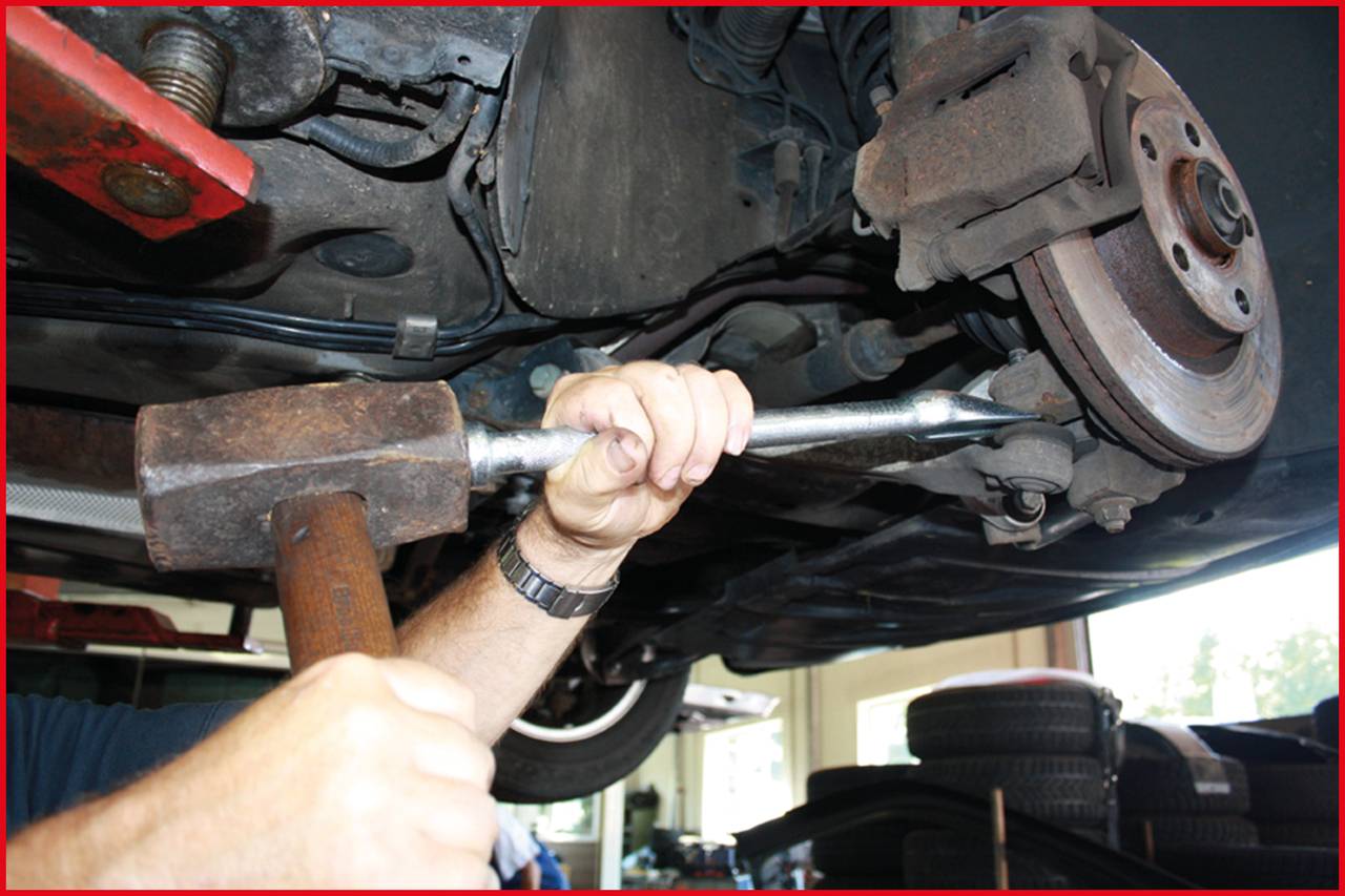 Nahaufnahme eines Mechanikers, der mit einem Hammer an einem Auto arbeitet. Die Aufnahme zeigt den Unterboden und ein Rad des Fahrzeugs.