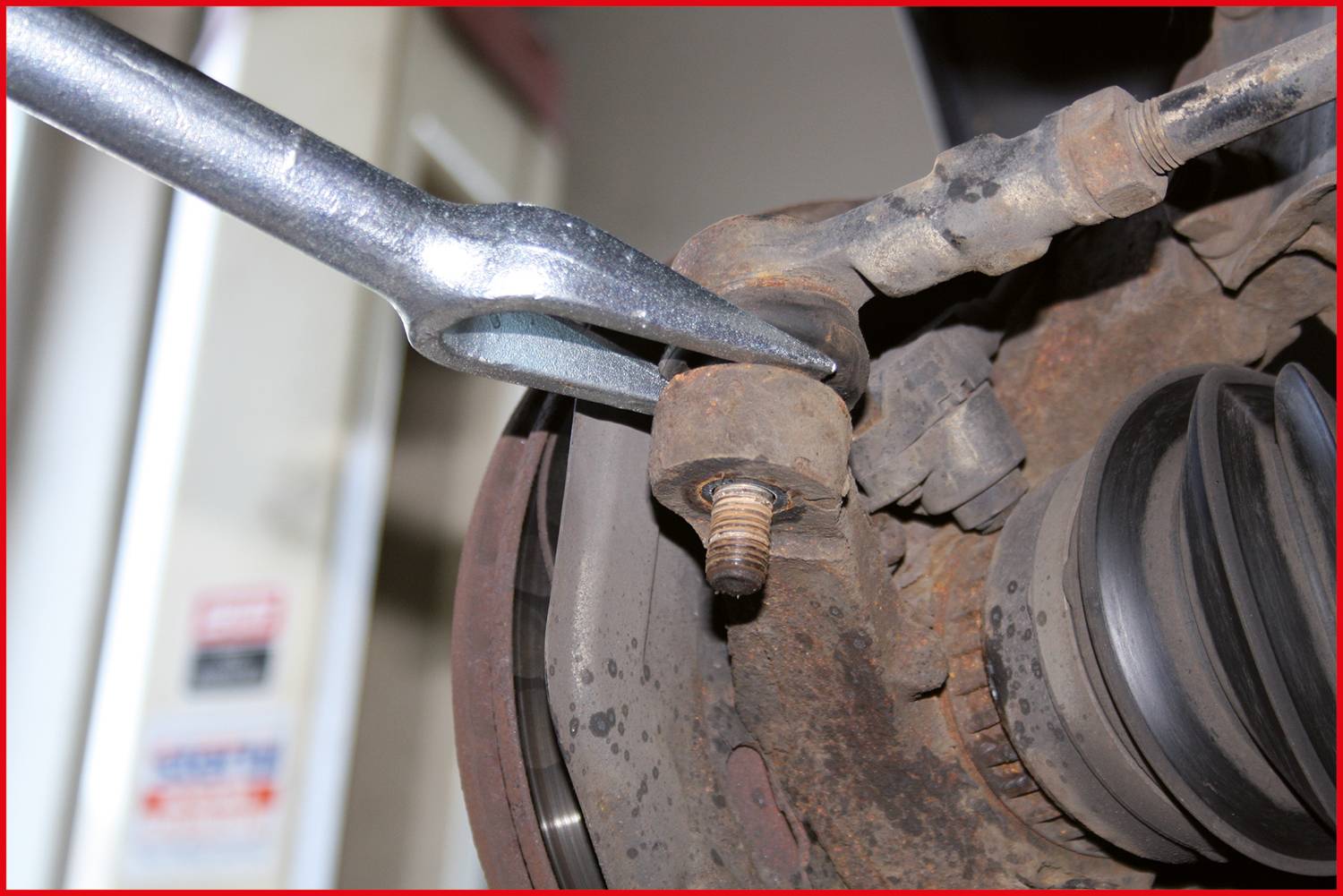 Mensch repariert Fahrwerksteil eines Fahrzeugs mit Metallwerkzeug in einer Werkstattumgebung. Fokus auf Schraubverbindung.
