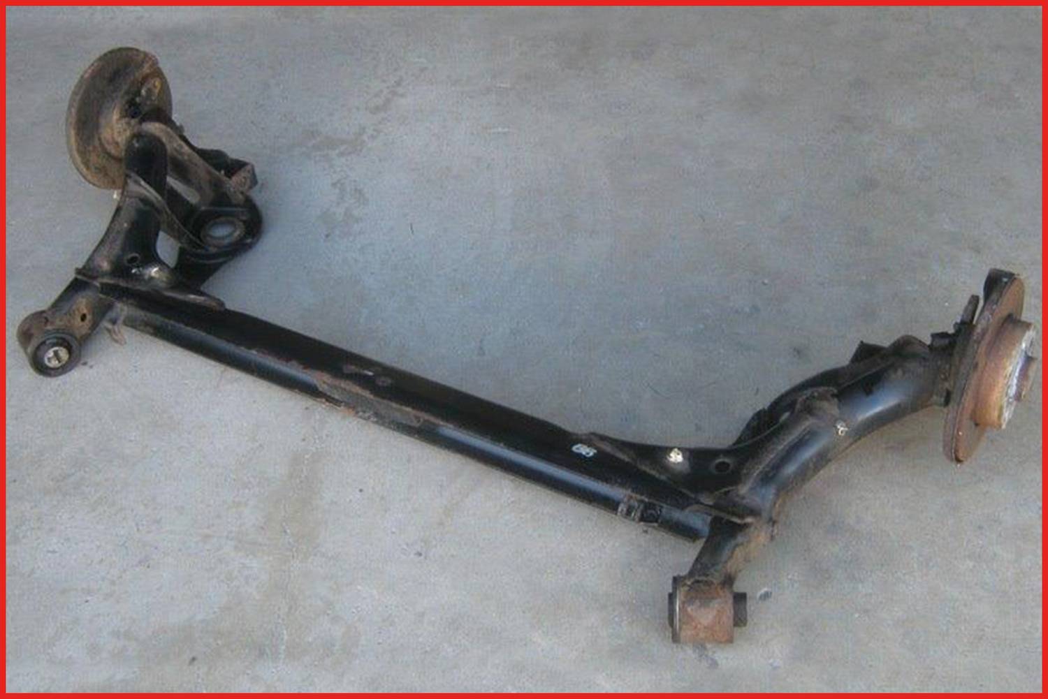 Eine schwarze Fahrzeug-Hinterachse liegt auf Betonboden. Sie zeigt zwei Radnaben an den Enden und Querstreben zur Verstärkung.