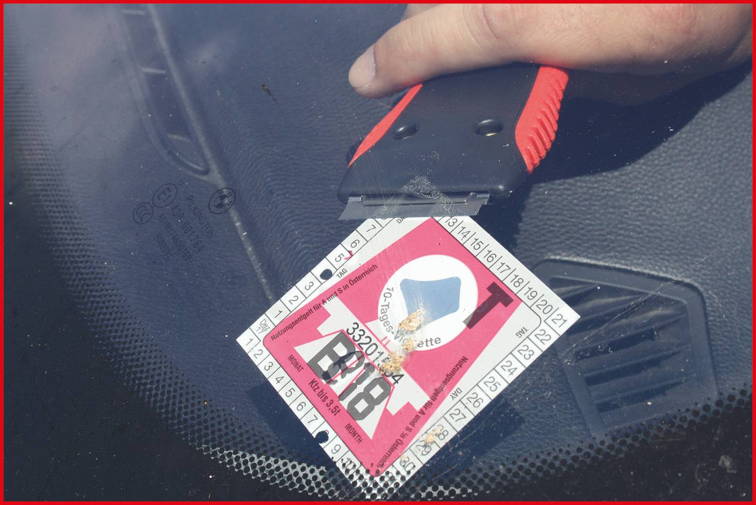 Hand verwendet einen Schaber, um eine Vignette von einer Windschutzscheibe zu entfernen. Die Vignette hat ein rot-weißes Design mit schwarzem Text.