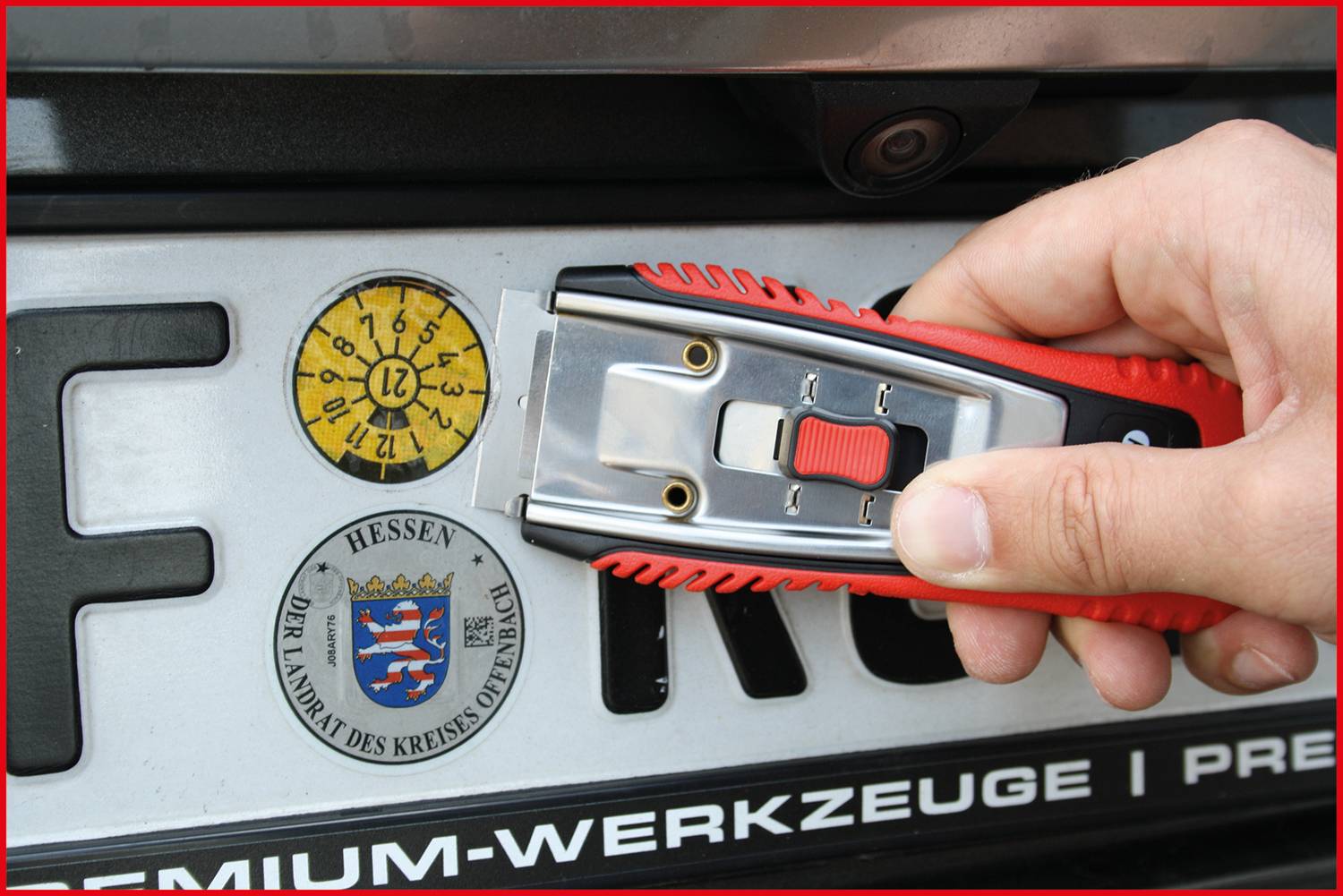Eine Hand hält einen Cutter über ein Kennzeichen mit Prüfplakette und Aufkleber des Kreises Offenbach, Hessen.