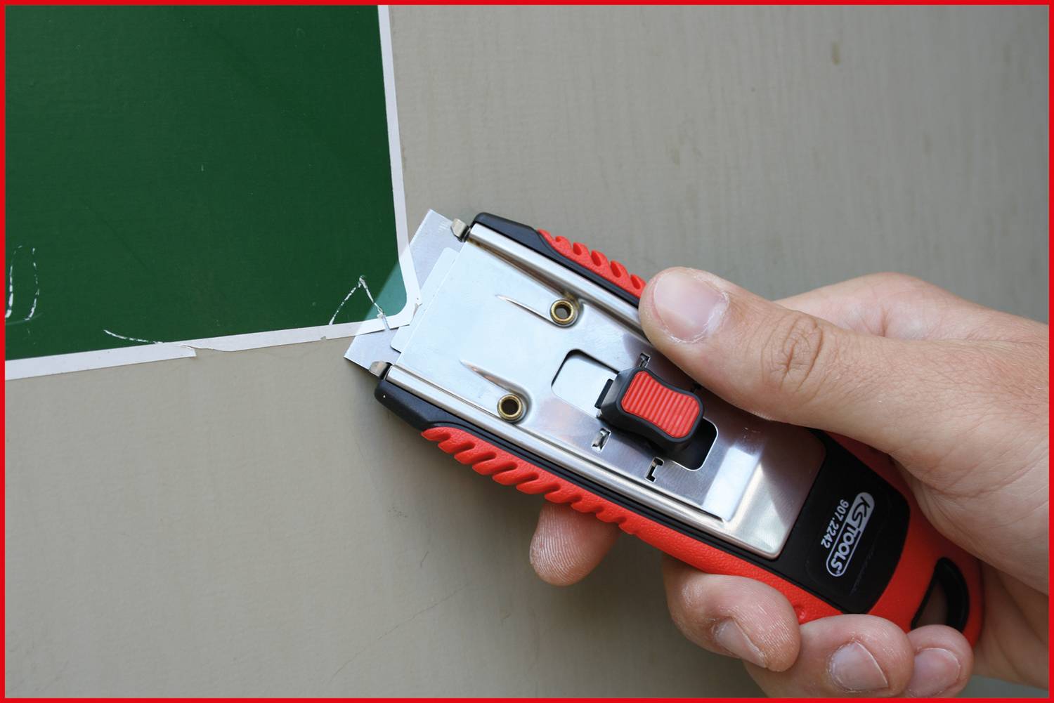 Hand hält einen roten Cutter mit schwarzem Griff und schneidet grüne Folie auf einer Oberfläche.