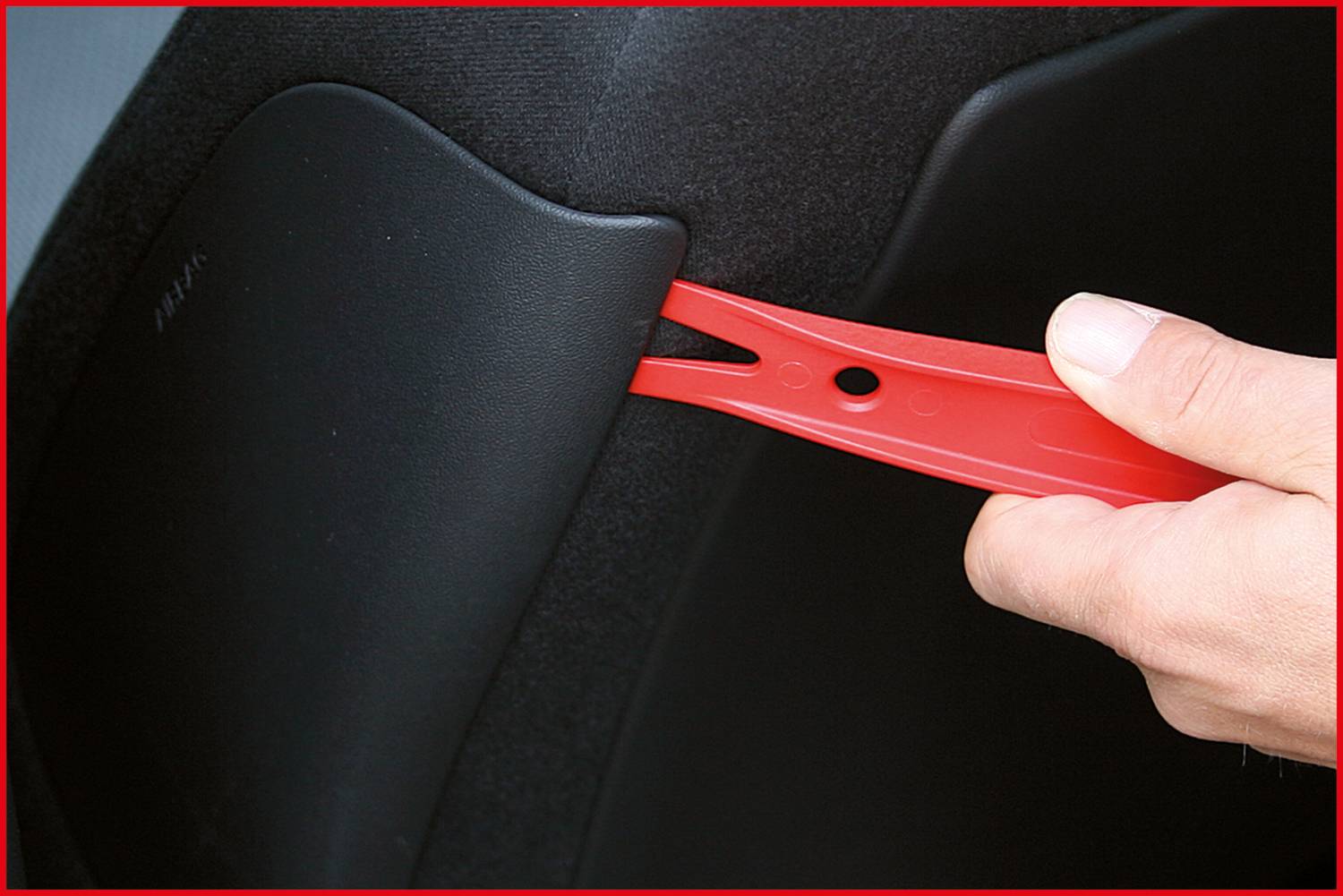 Ein Hand hält ein rotes Kunststoffwerkzeug, das hinter ein Autoteil geklemmt ist, vermutlich um es zu lösen oder zu entfernen.