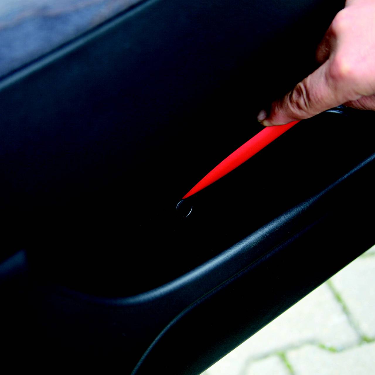 Eine Person hält ein rotes Objekt in einem schmalen Spalt eines Autoteils. Der Fokus liegt auf der Hand und dem Objekt.