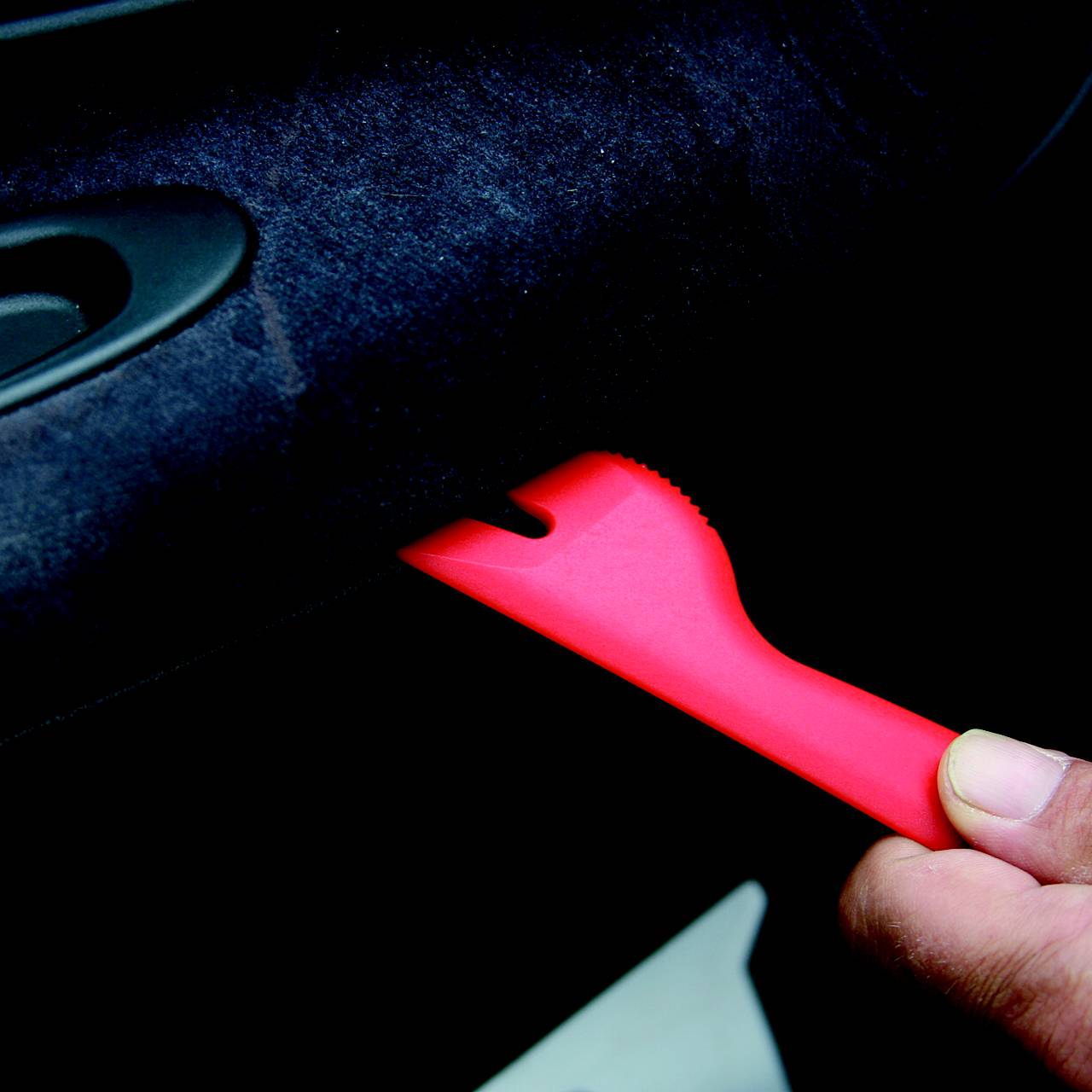 Eine Hand hält ein rotes Kunststoffwerkzeug, das in eine Spalte eines Fahrzeuginnenraums gesteckt wird, wahrscheinlich zur Demontage von Verkleidungen.