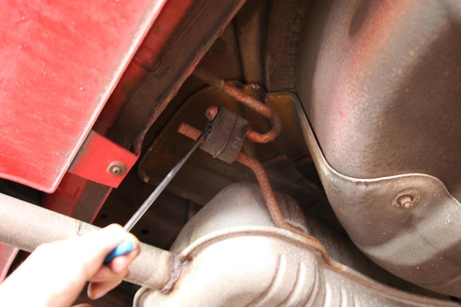 Eine Person repariert den Auspuff eines Autos mit einem Schraubenschlüssel. Man sieht den Unterboden und Teile des Auspuffsystems.