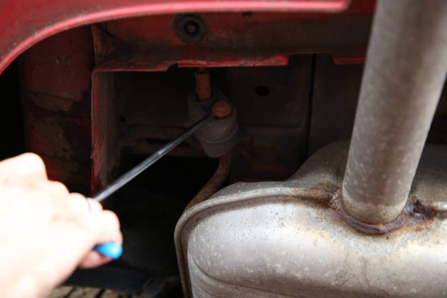 Person repariert Auspuff; Hand hält blauen Schraubenzieher, richtet ihn auf rostiges Teil. Fahrzeugunterboden im Hintergrund sichtbar.