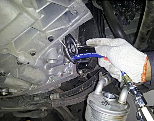 Ein Mechaniker repariert den unteren Teil eines Motors, eine Hand mit Handschuh hält ein Werkzeug. Im Vordergrund sind Rohre sichtbar.