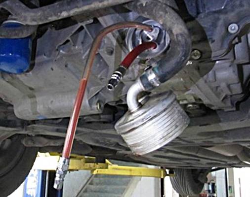 Ölablassvorgang bei einem Fahrzeug in einer Werkstatt. Ein Schlauch leitet das abgelassene Öl von der Unterseite des Motors in einen Behälter.