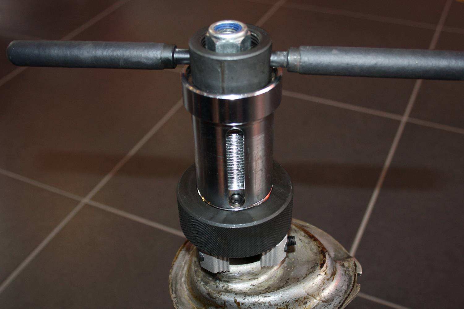 Nahaufnahme eines metallischen Werkzeugs mit Griffen und Gewinde, das auf einem Stativ montiert ist, vor einem gefliesten Hintergrund.