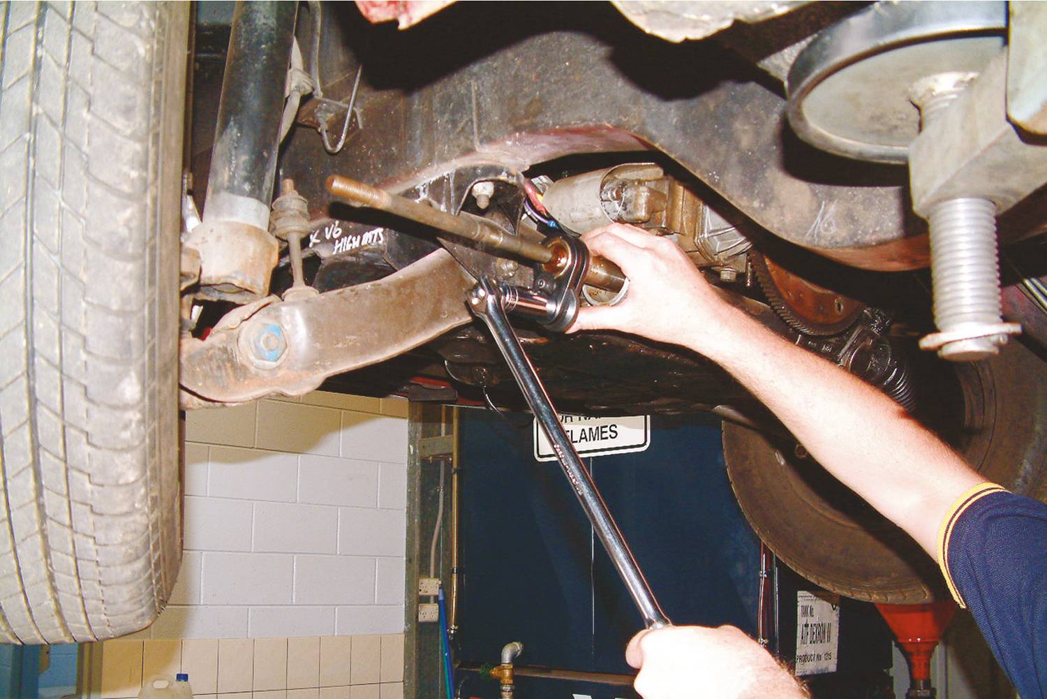 Eine Person repariert ein Auto von unten in einer Werkstatt. Sie verwendet einen Schraubenschlüssel, um an einer Fahrzeugachse oder einem Fahrwerkselement zu arbeiten.