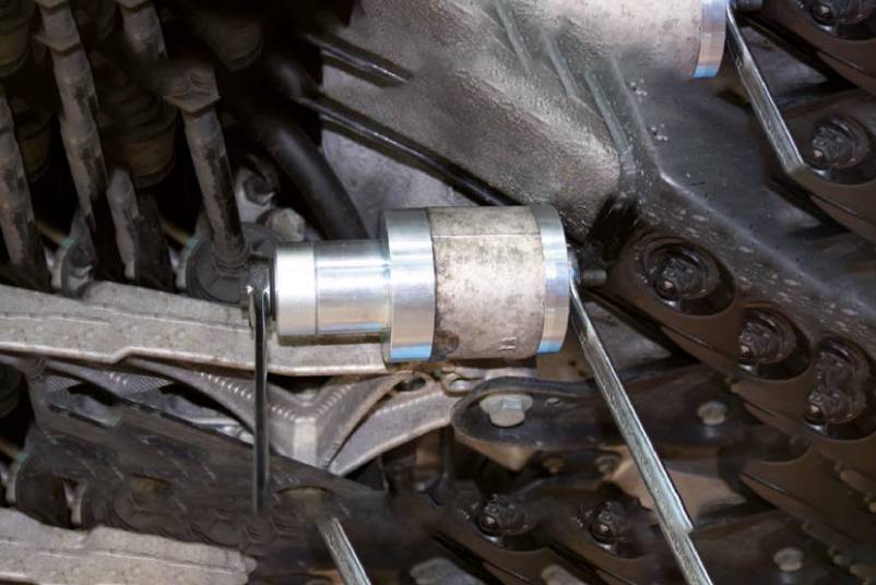 Ein Metallwerkzeug ist an einem Kettenantrieb in einem Motorraum befestigt. Es scheint zur Einstellung oder Wartung zu dienen.