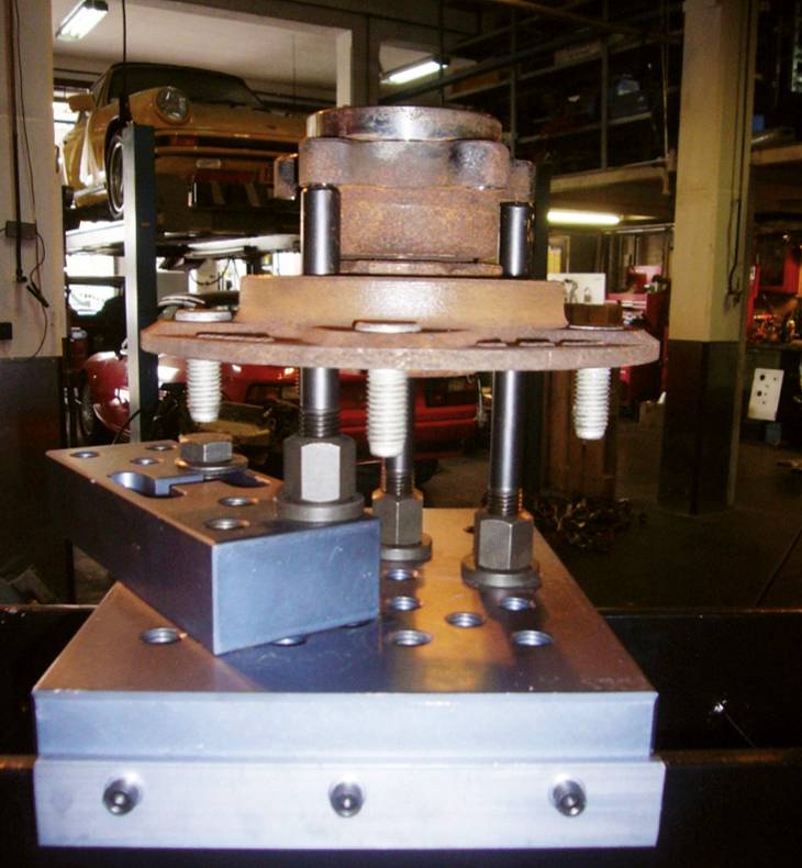 Stanzmaschine in einer Werkstatt, Schrauben und Metallplatten klemmen ein Werkstück. Im Hintergrund sind Autos auf Hebebühnen sichtbar.