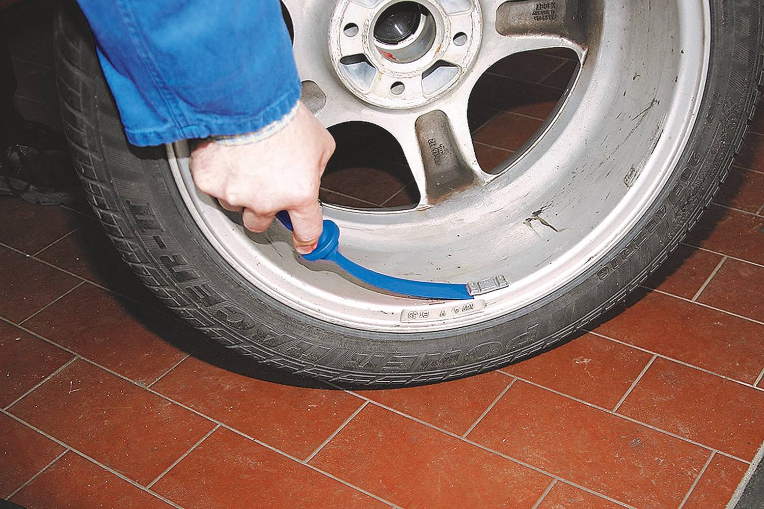 Eine Person misst mit einem blauen Werkzeug die Profiltiefe eines Autoreifens, der auf einem roten Fliesenboden liegt.