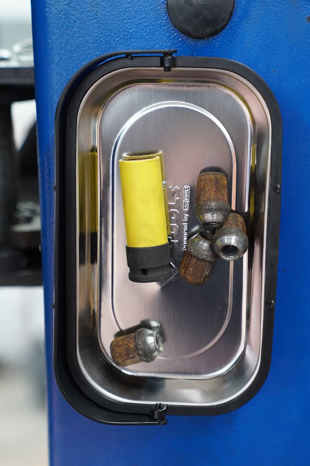 Ein metallener Behälter an einer blauen Maschine enthält einen gelben Adapter und mehrere rostige Schraubenmuttern.