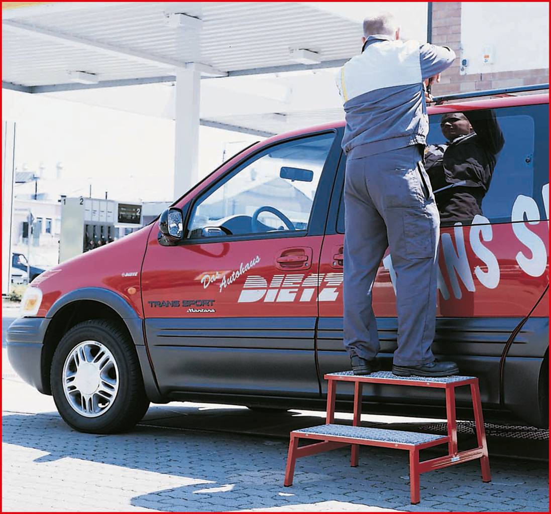 Ein Mann in Arbeitskleidung montiert einen Spiegel an einem roten Transporter bei einer Tankstelle. Eine Leiter steht neben dem Fahrzeug.