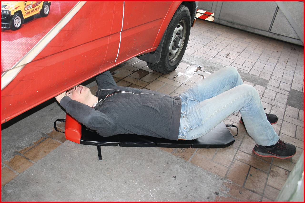 Eine Person liegt auf einem Rollbrett unter einem roten Lieferwagen, offensichtlich bei einer Reparaturarbeit in einer Garage.