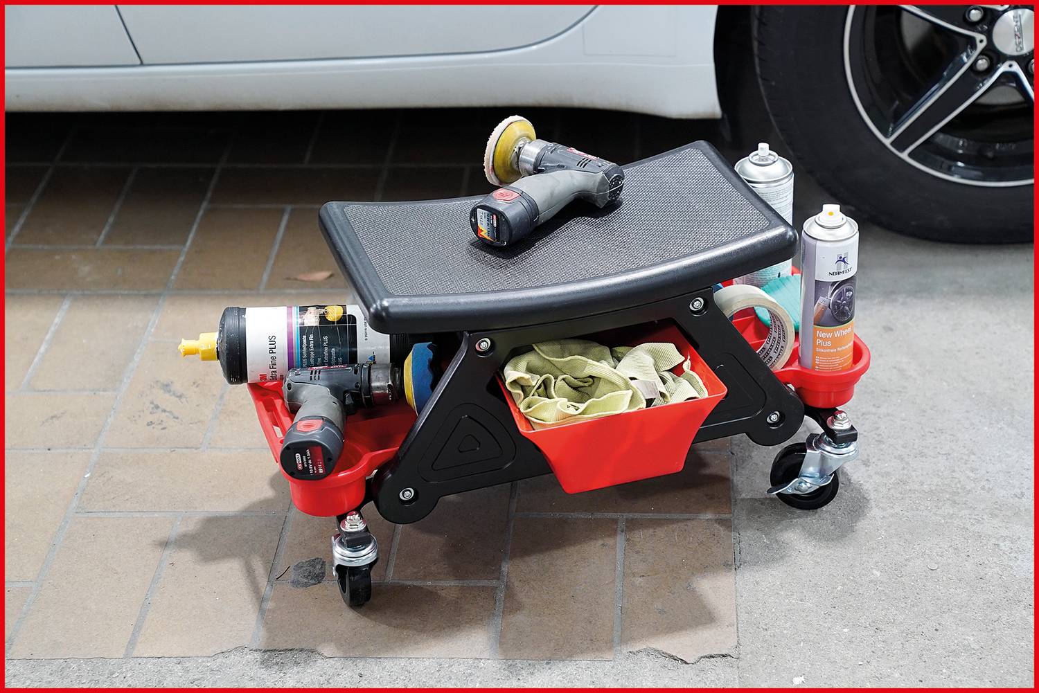 Ein mobiler Werkstattwagen mit Sitzfläche ist mit Poliermaschine, Reinigungsmitteln und Tüchern ausgestattet, steht neben einem Auto.