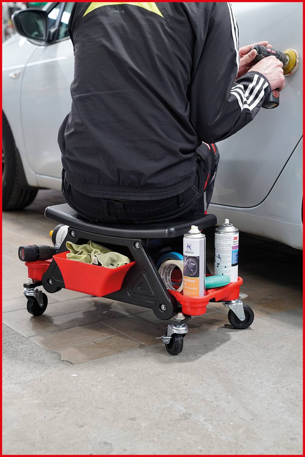 Person sitzt auf einem mobilen Werkstattrollhocker und poliert ein Auto, während Werkzeuge und Pflegeprodukte im Ablagefach sichtbar sind.