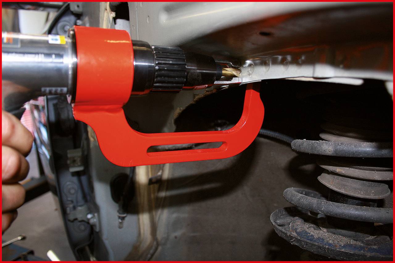 Nahaufnahme einer roten Metallklammer, die an einem Autoteil befestigt ist. Ein Bohrer wird verwendet, um ein Loch in das Blech zu bohren.